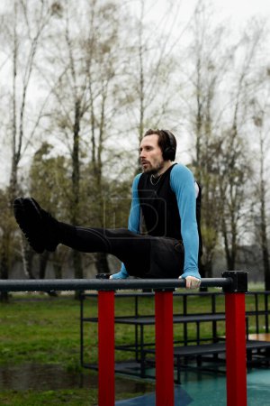 Téléchargez les photos : Sportsman équilibrage des jambes à l'aide de barres parallèles. Puissant jeune homme qui fait l assis abdos sur des poignées en métal. Entraînement de fitness sur terrain de sport. Exercice de gymnastique dans le parc. Photo de haute qualité - en image libre de droit