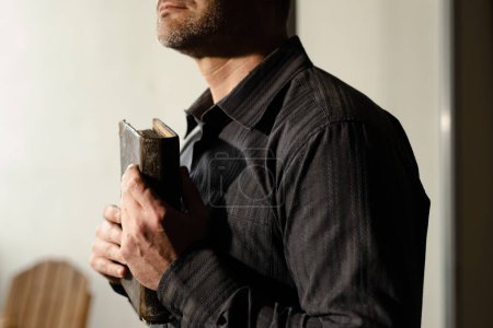 Photo for Close up shot of human in black clothing hugging holy bible. Mature man praying to god with hands on old scriptures. Male studying religion literature. Orthodox worship. High quality photo - Royalty Free Image