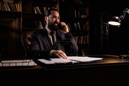 Photo for Ceo big corporation working at late nights. Positive businessman makes a phone call about some documents in the evening. Bearded man workaholic wear elegant formal suit and talking to smartphone. High - Royalty Free Image
