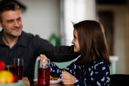 Photo for Concept of happy family. Mid shot proud father stroking his little daughter. Daddy listening his cute small girl at dining table. Christian father and schoolgirl at morning having meal. High quality - Royalty Free Image