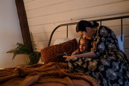 Foto de Concepto de familia feliz. Joven madre y su hija jugando videojuegos en la tableta. Mamá usa la computadora inalámbrica para mostrar la película de su hija a la hora de acostarse. Criar momentos de felicidad por la noche. Alto. - Imagen libre de derechos