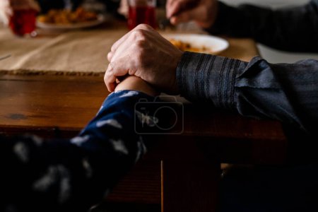Photo for Close up shot of family holding hands together at dining table. Christians praying before eating. Spiritual tradition clasping arms on the desk. Catholic church believers parents thanks god. High - Royalty Free Image