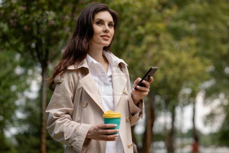 Photo for A brunette lady walks in the spring park, holding a cup of coffee and a smartphone in hands. An attractive girl stands outdoors with a slight smile on her face, waiting for an schedule appointment or - Royalty Free Image