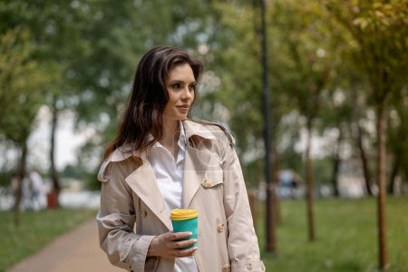 Téléchargez les photos : Une jeune femme se promène dans le parc printanier avec une tasse de café après une journée de travail et profite de la nature à l'extérieur. Une jolie brune se tient sur le côté et regarde la distance pensivement. Un spectaculaire - en image libre de droit