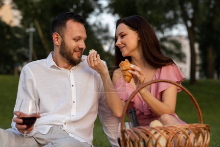 Photo for Cute young girlfriend feeds her boyfriend french yummy croissant on picnic. Couple having romantic date outdoors. Wife giving husband a small piece of bakery in park. Family vacation. High quality - Royalty Free Image