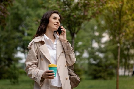 Photo for A positive smiling lady uses smartphone, has a pleasant conversation with a friend, boyfriend or colleague. A pretty young woman walks in a spring park with a cup of coffee outdoors. An attractive - Royalty Free Image