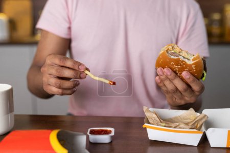 Photo for A young african or american man in the kitchen holds a hamburger and fries in hands, dipping potato in ketchup or sauce. A guy enjoys a snack of fast food in a cozy home environment. The selective - Royalty Free Image