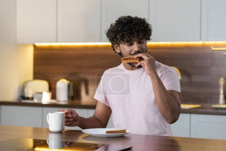 Téléchargez les photos : Un gars métis se tient dans la cuisine dans un éclairage tamisé, tient une tasse à la main, mange un sandwich et boit du thé ou du café les yeux fermés dans le plaisir. Un jeune homme américain ou africain déguste une collation - en image libre de droit