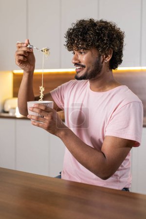 Téléchargez les photos : Un jeune homme métis souriant se tient sur le côté dans la cuisine dans un éclairage tamisé, tient un bol de pâtes dans les mains et les enveloppe dans une fourchette ludique. Un Américain positif ou un Africain apprécie un dîner - en image libre de droit