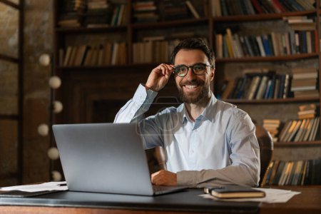 Foto de Un hombre sonriente ajusta las gafas con elegancia, se sienta en una mesa en la biblioteca y usa una computadora portátil. A un joven científico feliz se le ocurre un nuevo descubrimiento científico. Un hombre de negocios alegre gana una licitación para - Imagen libre de derechos