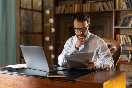 Photo for A man with serious look holds and reads important documents attentively, sits at a table in the library and uses a laptop. A respectable businessman acquainted with a lucrative contract. A young - Royalty Free Image