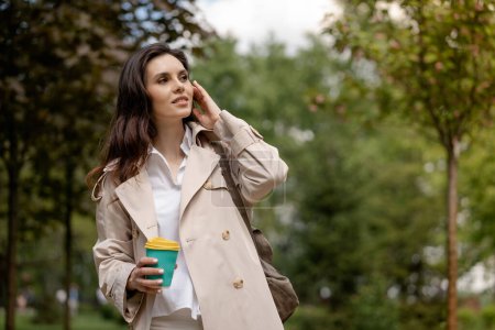 Téléchargez les photos : Une jolie jeune femme se relaxe lors d'une promenade dans un parc printanier avec une tasse de café à l'extérieur. Une dame élégante regarde vers l'avant avec un regard attentif, voir une personne familière, une vue intéressante ou - en image libre de droit