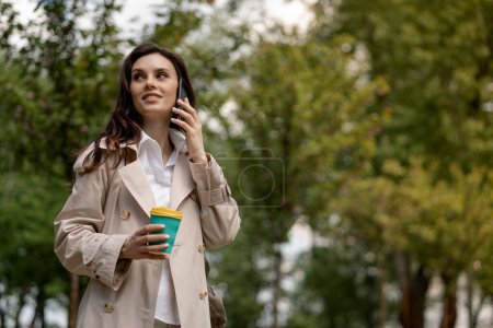 Photo for A smiling lady uses smartphone and has a pleasant conversation. A pretty young woman walks in a spring park with a cup of coffee outdoors. An attractive girl observes the surrounding nature and - Royalty Free Image