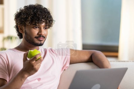 Photo for A mestizo guy with slight smile sits in the room on the sofa and enjoys relax after work or study in the cozy home environment. A young attractive african or american man eats apple, uses a laptop to - Royalty Free Image
