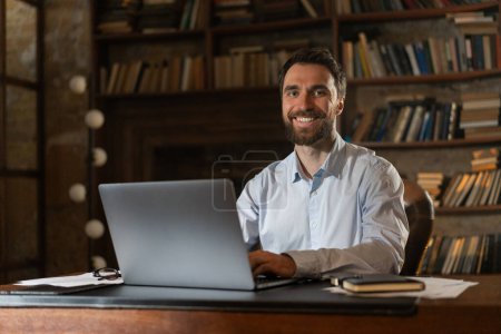 Téléchargez les photos : Un homme souriant s'assoit à une table de la bibliothèque et utilise un ordinateur portable. Un jeune scientifique se prépare à défendre candidat ou thèse de doctorat. Un homme d'affaires joyeux reçoit une offre pour rentable - en image libre de droit