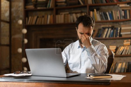 Photo for A man squints eyes and covers face with hand tiredly, sits at a table in the library and uses a laptop. A respectable businessman thinks hard about the upcoming important meeting. A young professor - Royalty Free Image