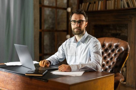 Photo for A respectable confident man sits at a table in the office library and uses a laptop. A young scientist prepares to defend dissertation. A businessman awaits the signing of a profitable international - Royalty Free Image