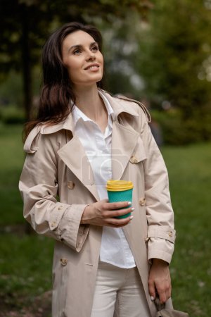 Téléchargez les photos : Une charmante jeune fille se détend dans un parc de printemps à l'extérieur avec une tasse de café après l'étude ou le travail. Un modèle de dame positive souriante dans un look coûteux pose en pleine hauteur pour des instantanés de - en image libre de droit