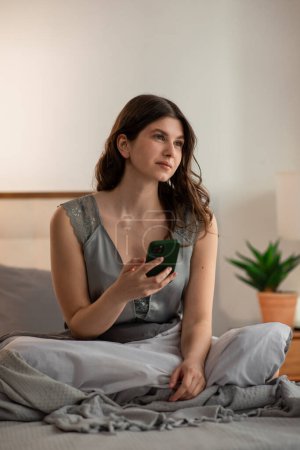 Photo for Cheerful young woman in sitting in bed with smartphone in the morning. Brunette girl is waiting for the call. High quality photo - Royalty Free Image