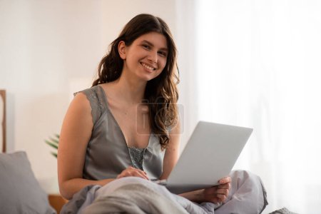 Photo for Smiling girl with laptop in bed looking at camera. Young woman typing on computer keyboard after awakening. High quality photo - Royalty Free Image