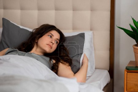 Téléchargez les photos : Brunette fille couchée sur l'oreiller après le réveil. Femme endormie au lit regardant l'alarme du téléphone tôt le matin. Photo de haute qualité - en image libre de droit