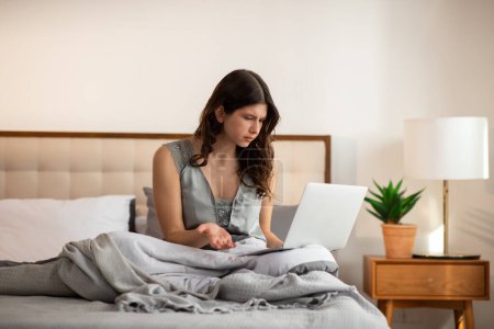 Photo for Irritated girl with laptop in bed is doing an urgent work. Working from home idea. Young woman typing on computer keyboard after awakening. High quality photo - Royalty Free Image