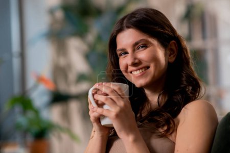 Foto de Hermosa chica morena está sosteniendo la taza de café y sonriendo a la cámara sobre un fondo borroso. Foto de alta calidad - Imagen libre de derechos