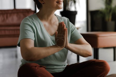Téléchargez les photos : Une femme d'âge moyen pratique le yoga à la maison. Une femme sans visage s'assoit latéralement sur le sol en position lotus avec les paumes de la main, pliées ensemble sur la poitrine et médite. Ferme là. Concentration sélective - en image libre de droit
