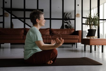 Téléchargez les photos : Une dame d'âge moyen pratique le yoga dans le grand hall de la maison. Une femme aux yeux fermés s'assoit détendue latéralement sur un tapis sur le sol en position lotus. Le concept de développement spirituel - en image libre de droit