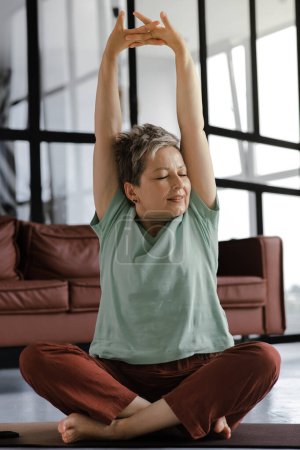 Téléchargez les photos : Une dame d'âge moyen pratique le yoga à la maison. Une femme avec les yeux fermés dans la félicité s'assoit sur un tapis sur le sol en position lotus, étire les bras au-dessus de la tête avec les paumes, face au plafond, et les doigts - en image libre de droit