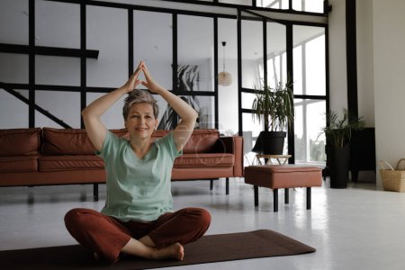 Photo for A middle aged lady practices yoga in the large hall of the house. A smiling woman sits on a mat on the floor in the lotus position and shows namaste gesture with palms, raised above head. Looks at the - Royalty Free Image