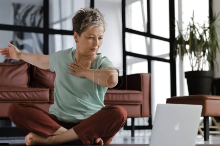 Foto de Una señora de mediana edad practica yoga en casa. Una mujer con mirada concentrada se sienta en una alfombra en el suelo en la posición de loto y mueve los brazos a los lados, utiliza computadora portátil, observa ejercicios y los repite - Imagen libre de derechos