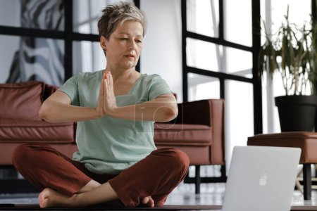 Téléchargez les photos : Une dame d'âge moyen pratique le yoga à la maison. Une femme au regard attentif s'assoit sur un tapis sur le sol en position lotus et paumes pliées ensemble sur la poitrine, utilise un ordinateur portable, regarde des exercices et répète - en image libre de droit