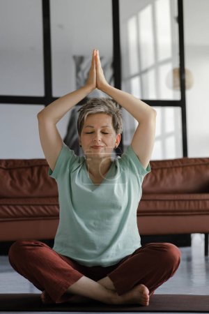 Téléchargez les photos : Une dame d'âge moyen pratique le yoga à la maison. Une femme aux yeux fermés s'assoit sur un tapis sur le sol en position lotus et montre un geste namaste avec les paumes levées au-dessus de la tête. Ferme là. Haute qualité - en image libre de droit