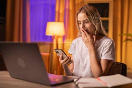 Photo for A girl student sits at a table in apartment, surrounded by laptop and copybook, uses smartphone and stunned by the unexpected news in the received message. A young lady demonstrates emotional stupor - Royalty Free Image
