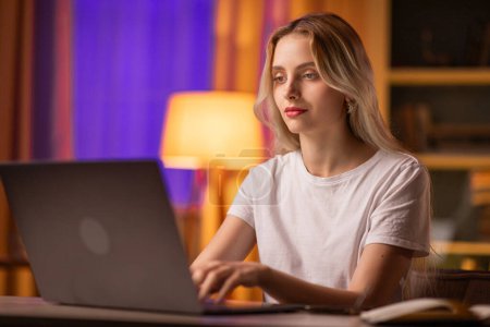 Photo for A girl student sits at a table in apartment, uses a laptop for typing a coursework or dissertation. A young businesswoman checks the profitability of own start up company remotely. A lady designer - Royalty Free Image