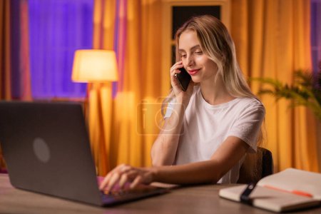 Photo for A girl student with a slight smile sits at a table in apartment, uses a laptop to search for educational materials and communicates on a mobile phone with a fellow student. A young lady sales manager - Royalty Free Image