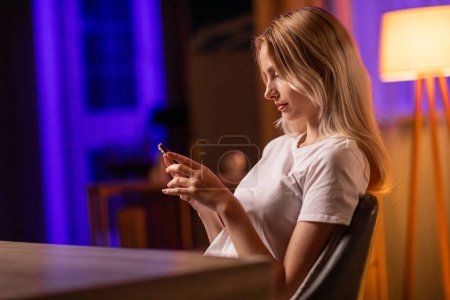 Photo for An attentive girl student sits sideways at a table in apartment, rests after studying and uses smartphone to view news feed on social network. A young lady reads an interesting ebook or watches a - Royalty Free Image