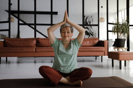 Foto de Una señora de mediana edad practica yoga en el gran salón de la casa. Una mujer sonriente se sienta en una alfombra en el suelo en la posición de loto y muestra gesto namaste con las palmas, levantado por encima de la cabeza. Mira el - Imagen libre de derechos