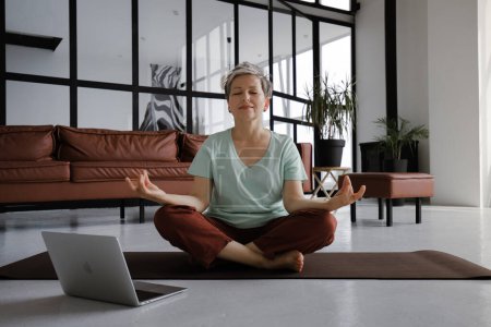 Foto de Una señora de mediana edad está practicando yoga en casa. Una mujer con una leve sonrisa en la cara se sienta en una alfombra en el suelo en la posición de loto, utiliza un ordenador portátil para ver videos relajantes y escuchar meditativo - Imagen libre de derechos