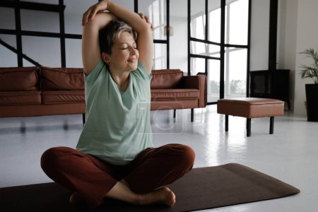 Téléchargez les photos : Une dame d'âge moyen pratique le yoga à la maison. Une femme souriante avec les yeux fermés dans la félicité s'assoit sur un tapis sur le sol dans la position du lotus et plie les bras au-dessus de la tête. Le concept de trouver l'harmonie du corps - en image libre de droit