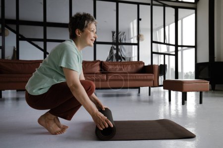 Foto de Una señora de mediana edad practica yoga en casa. Una mujer sonriente en un estado de ánimo satisfecho se agacha de lado y descalza de puntillas, rueda la esterilla después del entrenamiento. De cerca. Mira la cámara. Alta calidad - Imagen libre de derechos