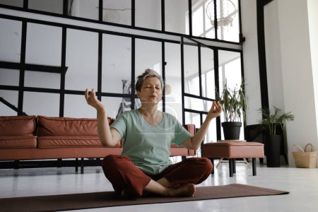 Téléchargez les photos : Une femme d'âge moyen pratique le yoga à la maison. Une femme aux yeux fermés et souriant sur un visage s'assoit détendue sur un tapis sur le sol en position lotus. Le concept de développement spirituel à travers - en image libre de droit