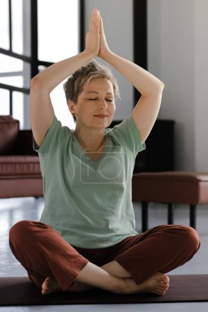 Foto de Una señora de mediana edad practica yoga en casa. Una mujer con los ojos cerrados se sienta en una alfombra en el suelo en la posición de loto y muestra gesto namaste con las palmas, elevado por encima de la cabeza. De cerca. Alta calidad - Imagen libre de derechos