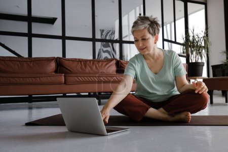 Foto de Una señora de mediana edad está practicando yoga en casa. Una mujer sonriente se sienta en una alfombra en el suelo en la posición de loto, tiene un tiempo de descanso del entrenamiento, utiliza un ordenador portátil y se ríe, mirando distracción divertida - Imagen libre de derechos