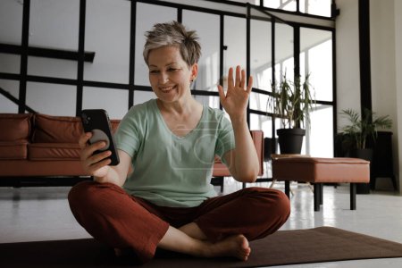 Téléchargez les photos : Une dame d'âge moyen pratique le yoga dans le grand hall de la maison. Une femme souriante est assise sur un tapis sur le sol en position lotus, a un temps de pause de la formation, utilise un smartphone pour la vidéo - en image libre de droit