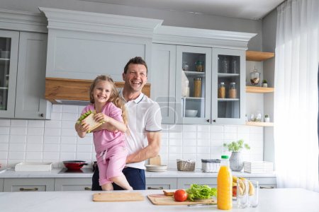 Foto de Una familia amigable de padre e hija sonrientes se paran en la cocina en un estado de ánimo alegre, se divierten, abrazan y ríen juntos. Un padre joven y un niño pequeño demuestran emociones positivas - Imagen libre de derechos