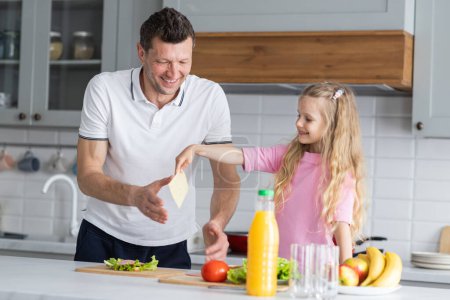 A friendly family from smiling father and daughter stand in the kitchen in a joyful mood, have fun, hug and laugh together. A young man dad and little girl child demonstrate positive emotions