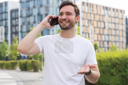 Téléchargez les photos : Un jeune homme souriant se tient dans la rue dans une ville du parc, utilise un smartphone et gesticule à la main. Un sportif entrepreneur de bonne humeur communique avec les autorités municipales sur l'idée de - en image libre de droit