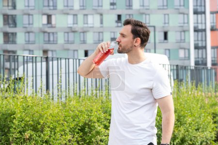 Photo for A young man has jogging or race walking in a modern residential area with colorful buildings and parkland on a summer morning. A sportsman stands sideways, takes a break in workout and drinks a - Royalty Free Image